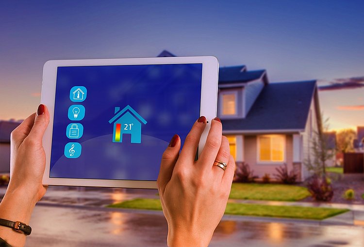 Mit Hilfe eines Tablets wird die Temperatur eines Hauses gesteuert, dass im Hintergrund zu sehen ist.