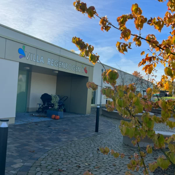 Eingangstür der Kinderkrippe Villa Regenbogen wird mit herbstlichen Bäumen gezeigt.